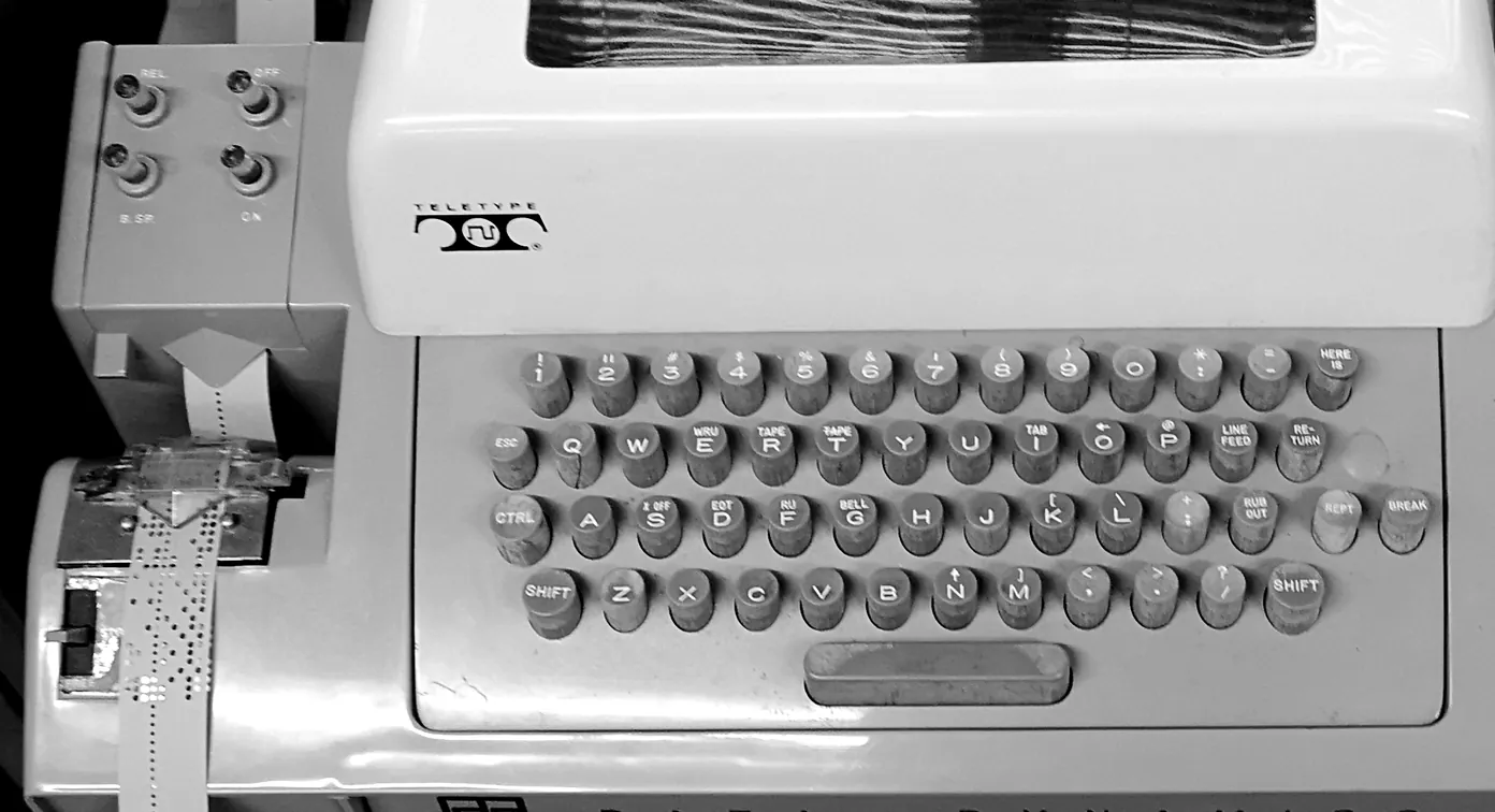 The left side of a Teletype Model 33 ASR. Four buttons on the tape punch are labelled REL., OFF, B.SP. and ON, and punched paper tape runs down from the punch. The keyboard to the right has a key marked RUB OUT.